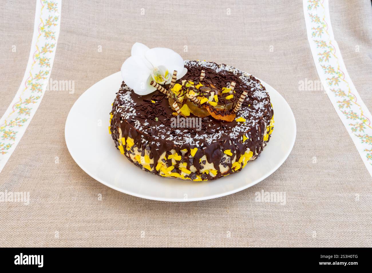 Un gâteau rond au chocolat est présenté sur une assiette blanche, orné de glaçage jaune et surmonté d’une fleur d’orchidée blanche, reposant sur une languette beige texturée Banque D'Images