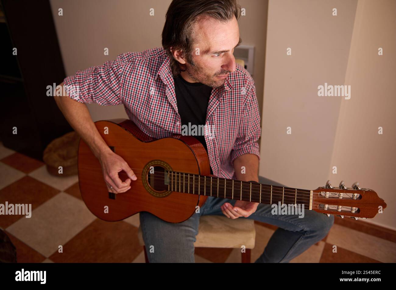 Un homme est assis à l'intérieur et joue de la guitare acoustique tout en étant concentré. Vêtu d'une tenue décontractée, il démontre des capacités musicales et un auto-expres artistique Banque D'Images