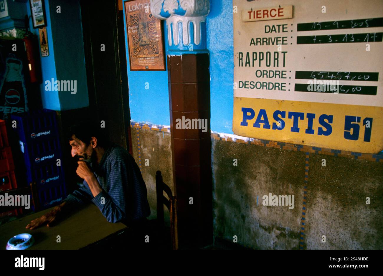 Un homme assis dans un café marocain à côté d'une planche Pastis 51 affichant les résultats des courses hippiques de l'époque coloniale française. Banque D'Images