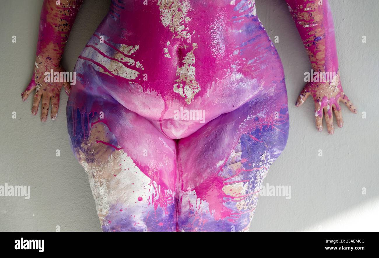 Un gros plan dynamique présente un corps de grande taille peint avec des éclaboussures de rose, violet et or, mêlant art et forme. Les mains expressives reposent sur un neutr Banque D'Images