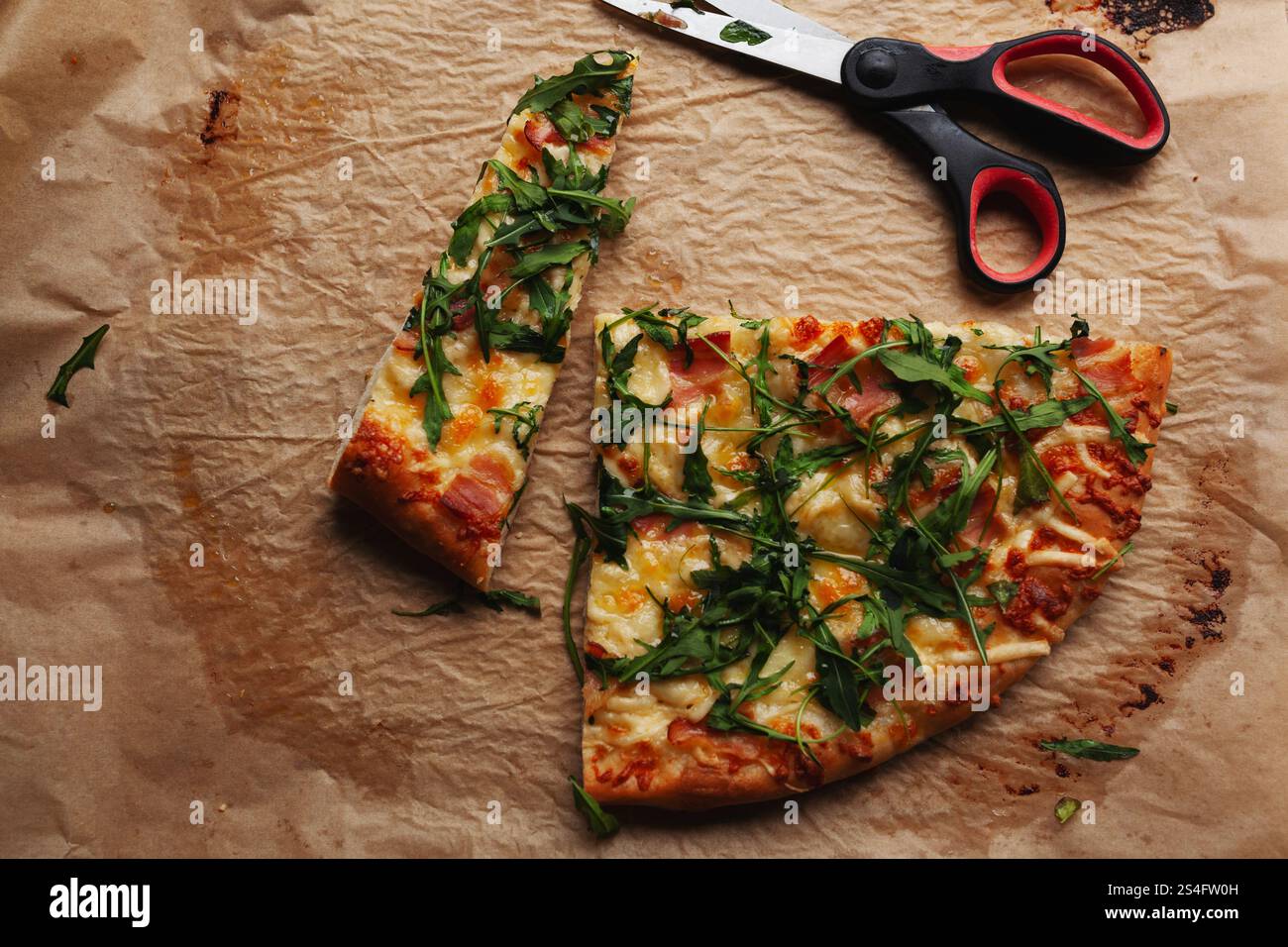 Pizza de style rustique sur du papier cuisson froissé, garnie de fromage fondu et de feuilles de roquette fraîches, une tranche coupée avec des ciseaux Banque D'Images