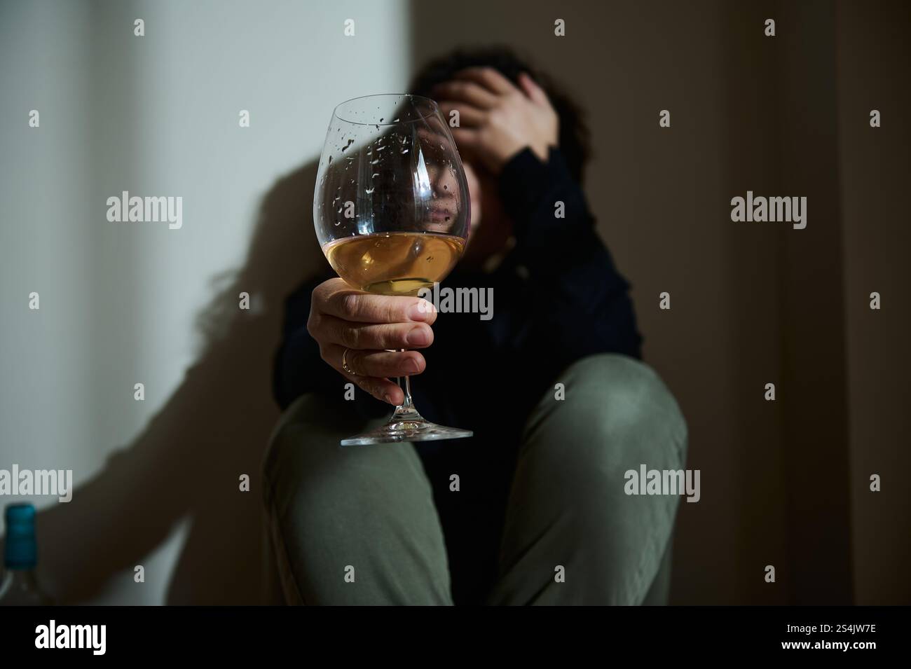 Une personne assise en basse lumière tient un verre de vin, symbolisant la détresse émotionnelle et l'auto-réflexion. Le cadre transmet des thèmes de luttes internes Banque D'Images