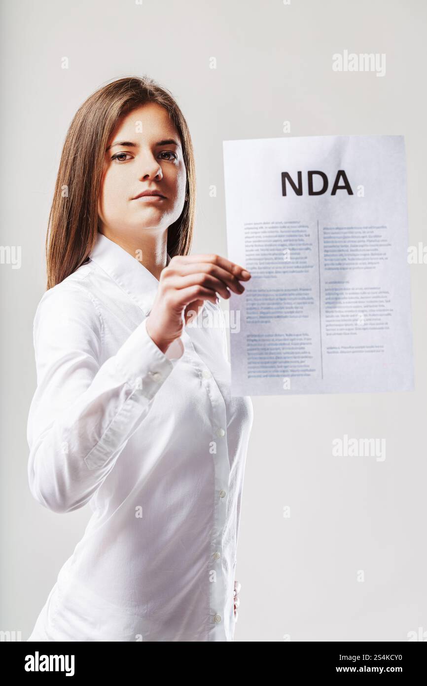 Jeune femme d'affaires détenant un document d'accord de non-divulgation (nda), soulignant la vie privée et la confidentialité avec une expression sérieuse contre un gr Banque D'Images