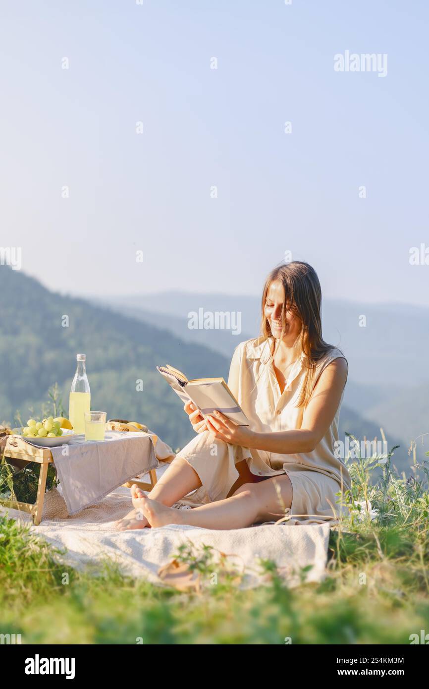 Femme profitant d'un pique-nique paisible dans les montagnes, lisant un livre et sirotant de la limonade par une journée ensoleillée. Banque D'Images