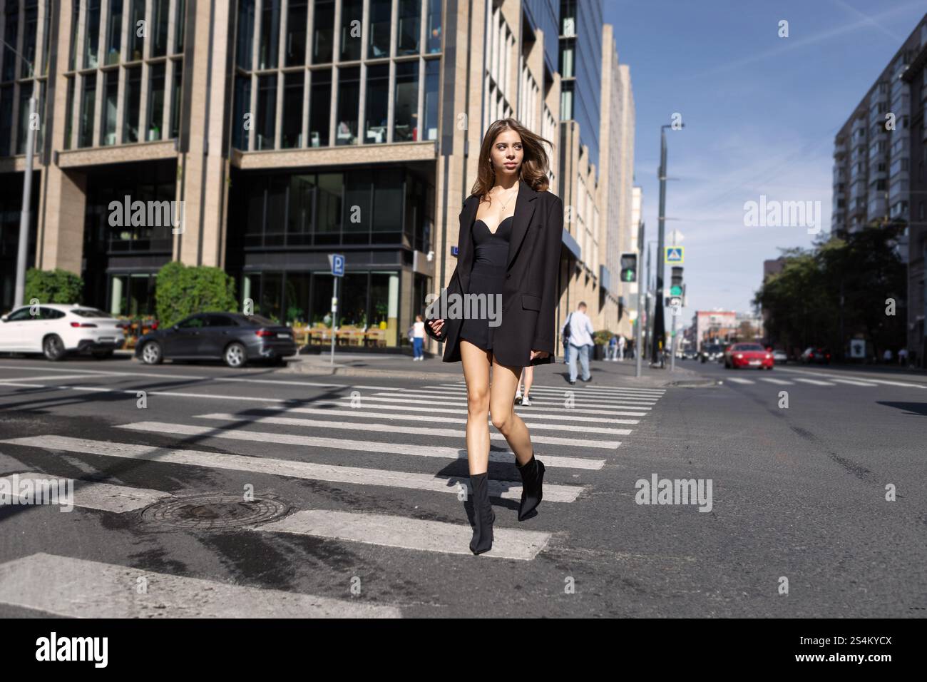 Élégante jeune femme de mode avec de longs cheveux marchant à travers le passage piétonnier dans la ville, portant une mini robe noire et des bottes, style urbain tendance M de rue Banque D'Images