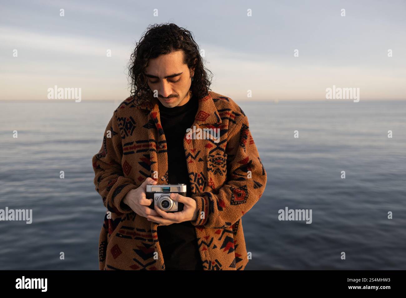 Un homme se tient près de la mer tranquille tenant un appareil photo vintage, enveloppé dans une veste à motifs le cadre serein capture un moment réfléchi contre un sof Banque D'Images