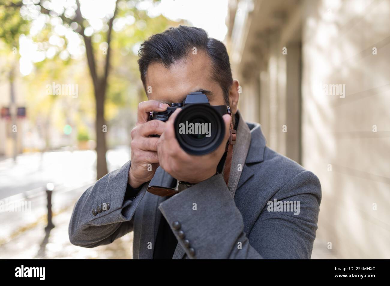 Un Indien, habillé comme un photographe avec un manteau gris, se concentre sur la capture d'images avec son appareil photo professionnel dans une ville verdoyante et ensoleillée Banque D'Images