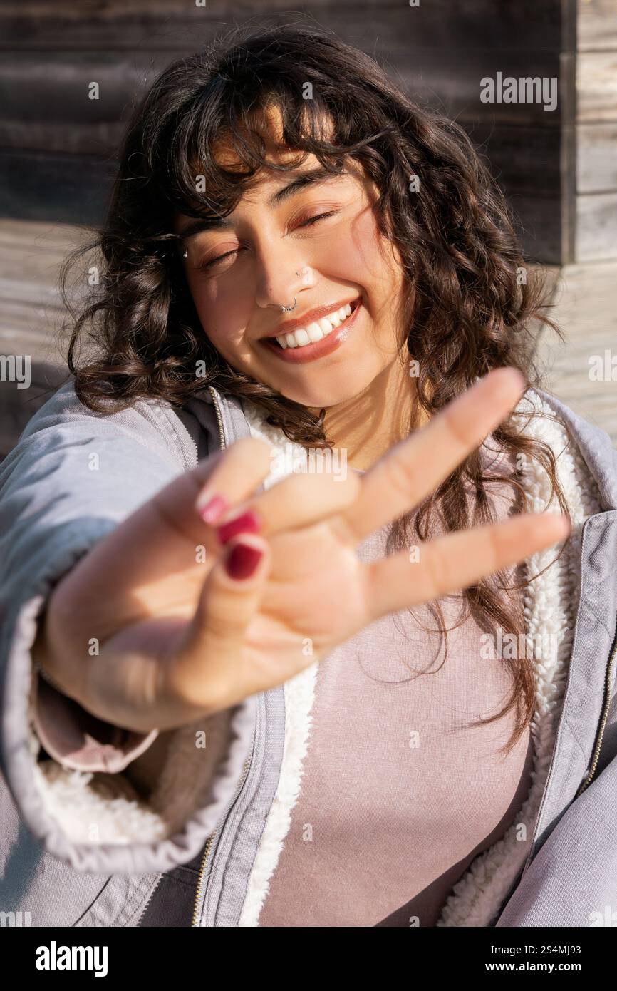 Une jeune femme latine dynamique profite d'une journée ensoleillée, clignotant un signe de paix avec un sourire joyeux. Son ambiance détendue et insouciante incarne le bonheur et la positivité Banque D'Images