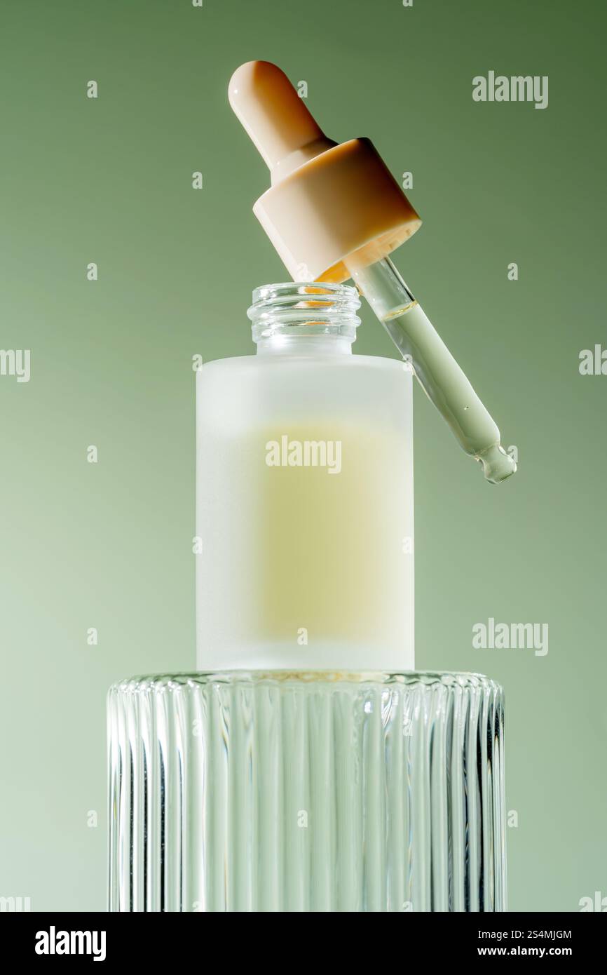 Un élégant flacon compte-gouttes de sérum avec une pipette reposant sur son bord, rempli d'un sérum laiteux, placé sur un fond vert doux. Banque D'Images