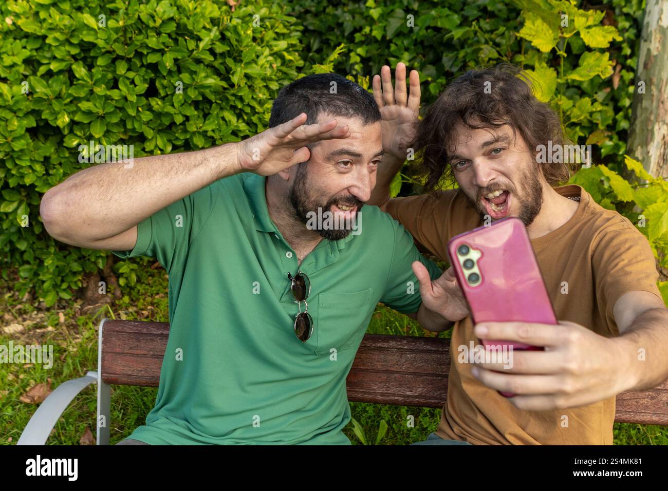 Deux amis s'amusent à prendre un selfie ludique assis sur un banc de parc l'homme en vert, qui est aveugle, sourit et pose avec enthousiasme aux côtés Banque D'Images