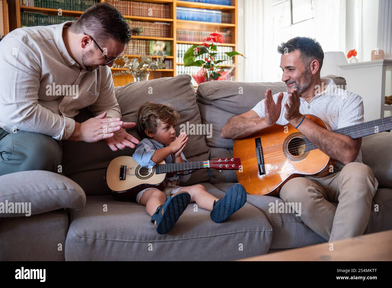 Un couple gay aimant profite du temps de qualité avec leur jeune fils, jouant de la guitare dans le salon confortable. Ils partagent des sourires et des applaudissements, encourageant la créat Banque D'Images