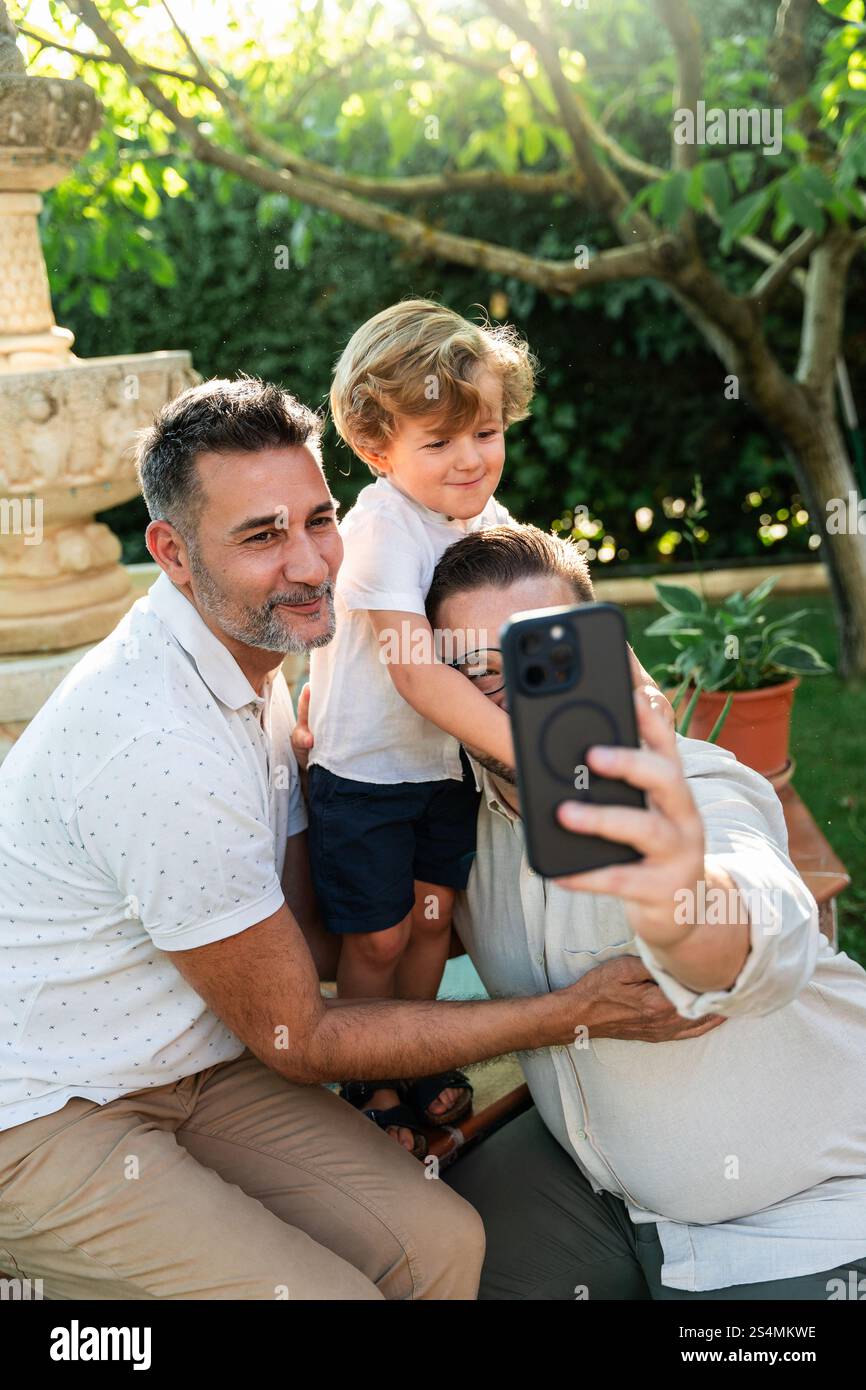 Un moment joyeux comme un couple gay prend un selfie avec leur fils dans un jardin ensoleillé. Embrasser la diversité et l'amour de la famille Banque D'Images Un moment joyeux comme un couple gay prend un selfie avec leur fils dans un jardin ensoleillé. Embrasser la diversité et l'amour de la famille Banque D'Images
