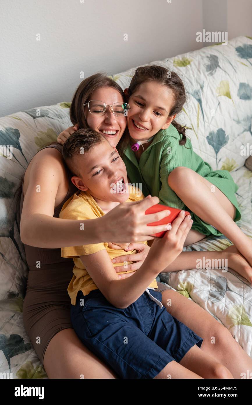 Une famille joyeuse partage un moment tendre sur le canapé. La mère aimante embrasse sa fille avec une trachéotomie et son fils avec une paralysie cérébrale, capturin Banque D'Images
