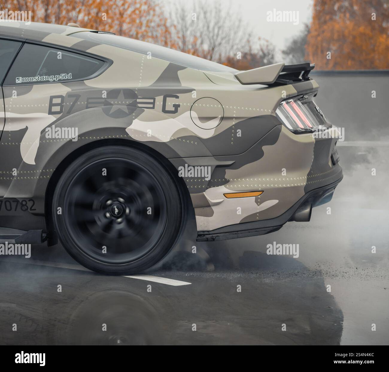 eige camo Ford Mustang faisant un burnout. Vue latérale arrière d'une voiture de poney américaine brûlant du caoutchouc sur une surface de béton humide. Banque D'Images