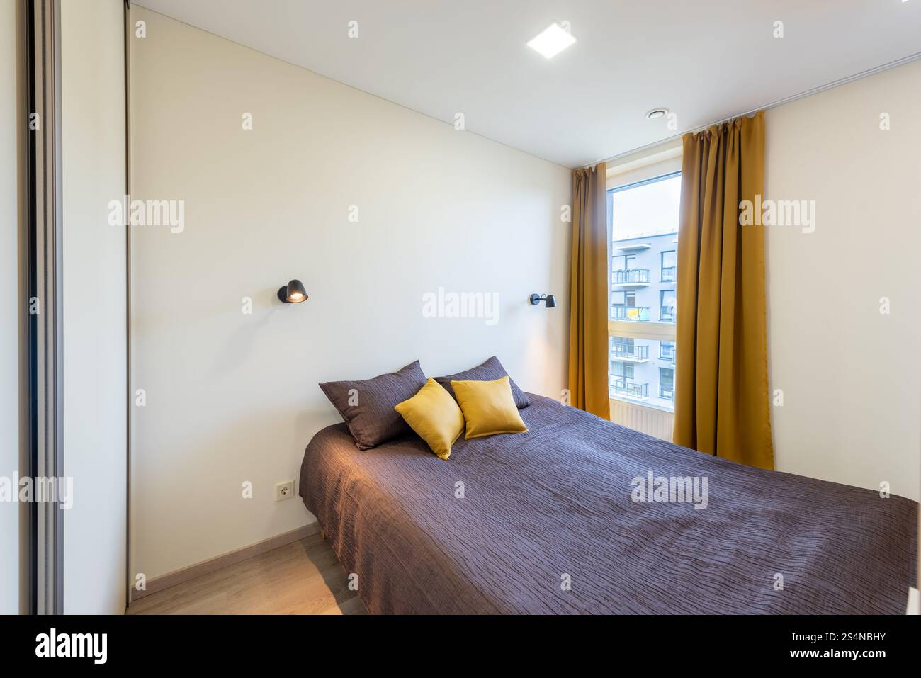 Cette chambre lumineuse et accueillante présente un décor élégant avec des rideaux moutarde, un lit confortable avec des coussins et un éclairage mural moderne. Lumière naturelle fi Banque D'Images