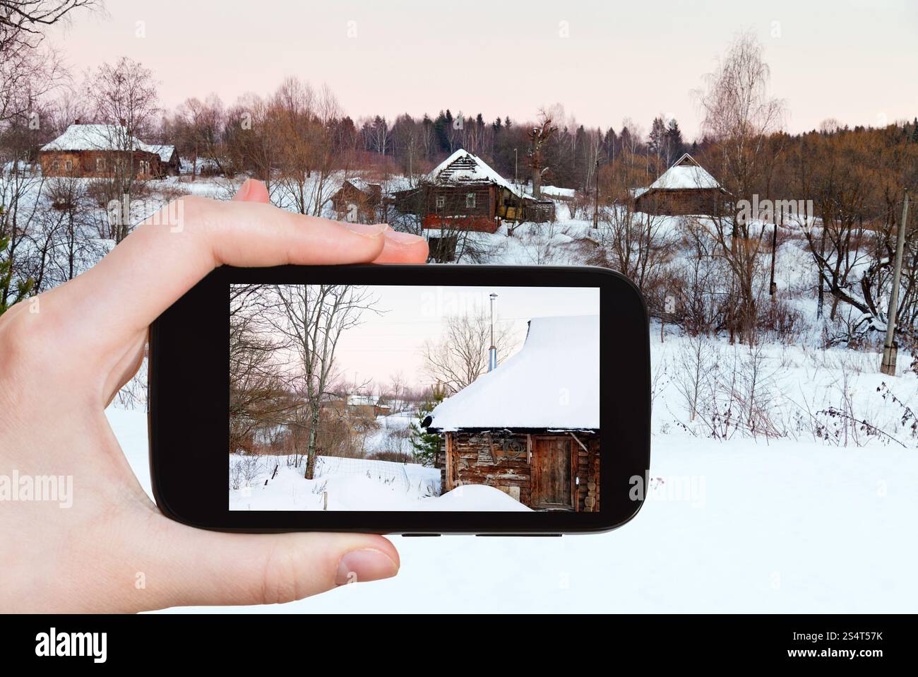 Concept de voyage - touriste prend des photos de paysage rural avec des maisons en bois enneigées dans le pays au coucher du soleil rose d'hiver sur smartphone, Russie Banque D'Images