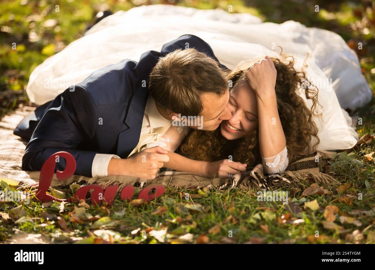 Beau jeune marié embrassant belle mariée dans la joue couchée sur l'herbe au parc Banque D'Images
