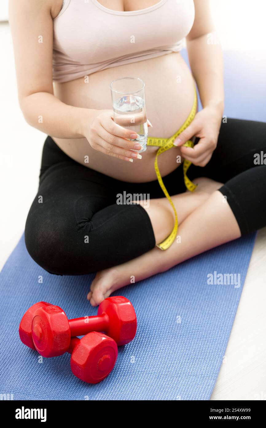 Pregnant woman measuring belly avec ruban à mesurer sur le tapis de fitness et holding glass of water Banque D'Images