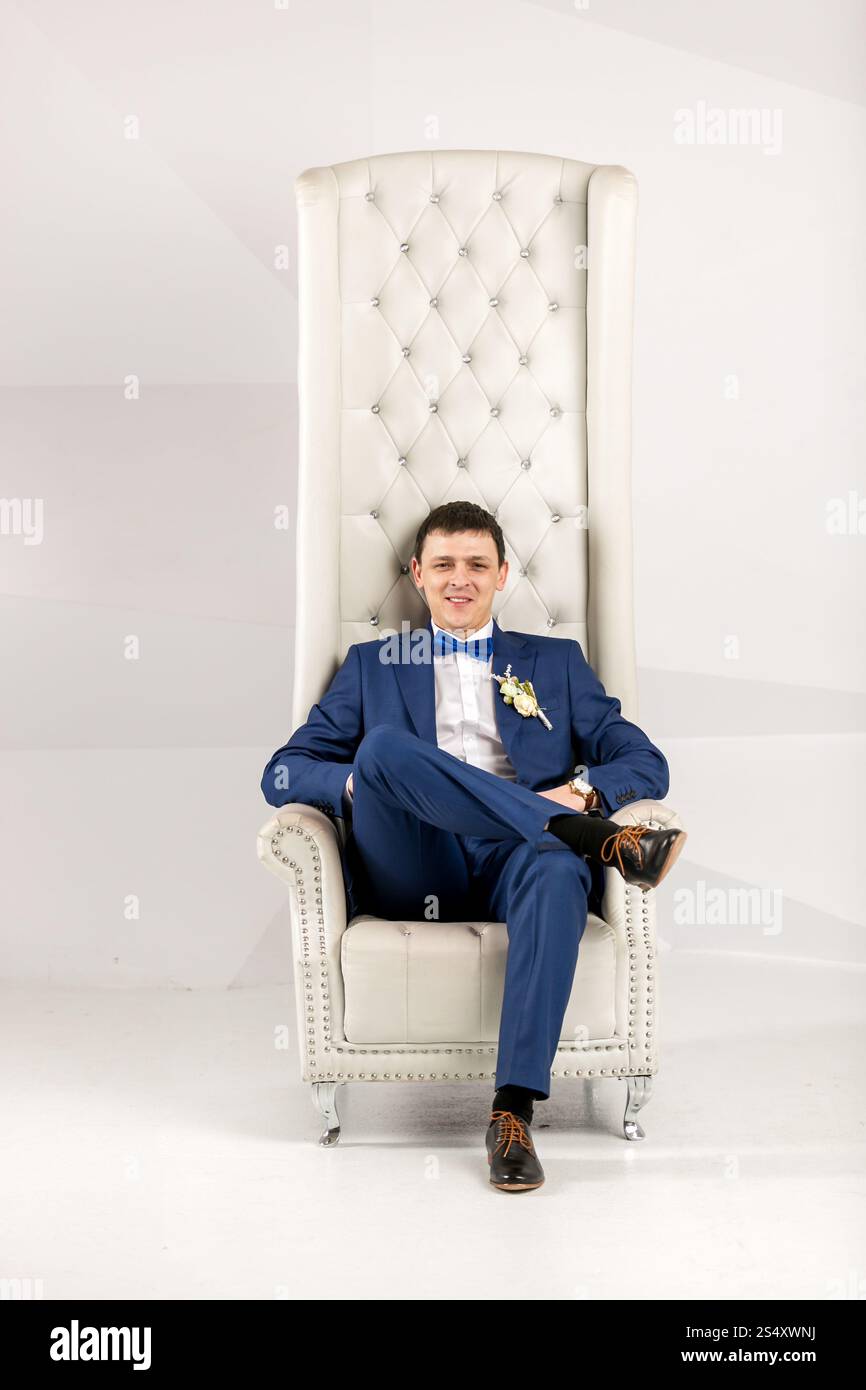 Bel homme élégant costume bleu en posant dans un fauteuil blanc au studio Banque D'Images