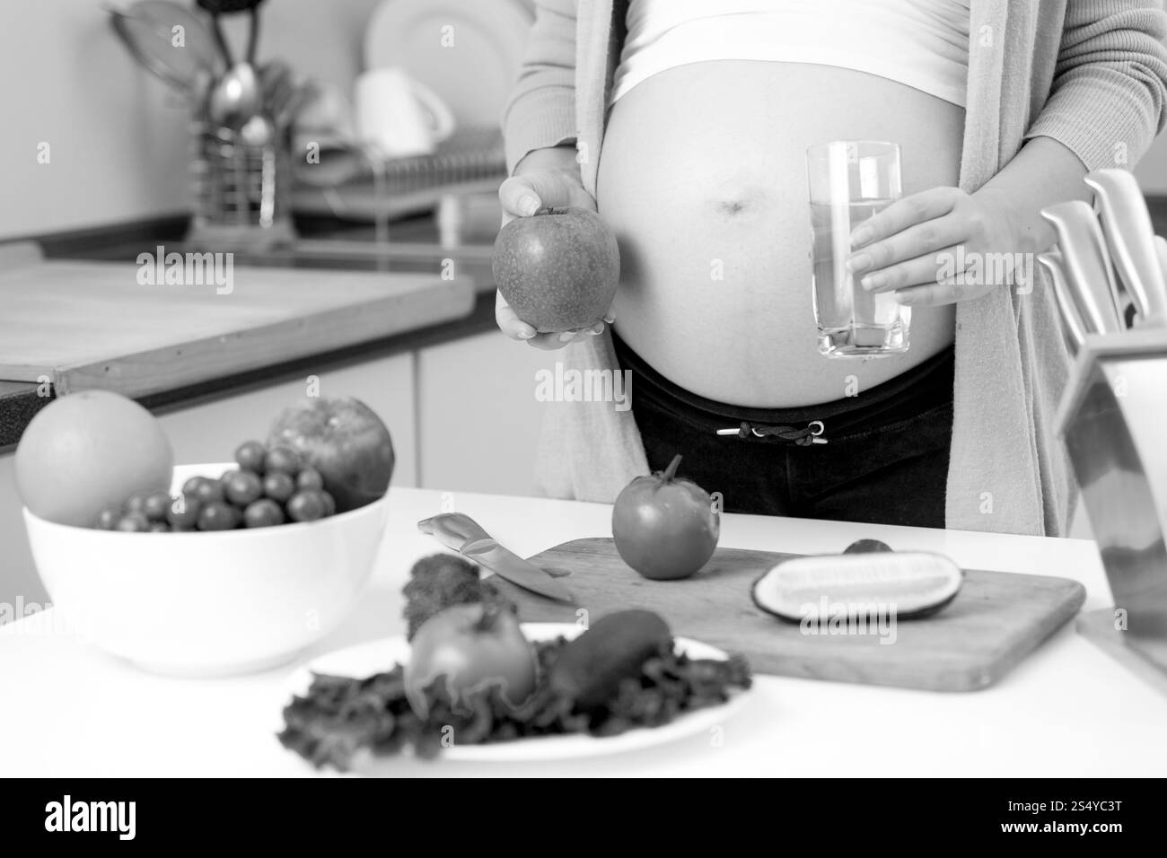 Gros plan photo en noir et blanc d'une femme enceinte posant sur la cuisine avec une pomme et un verre d'eau Banque D'Images