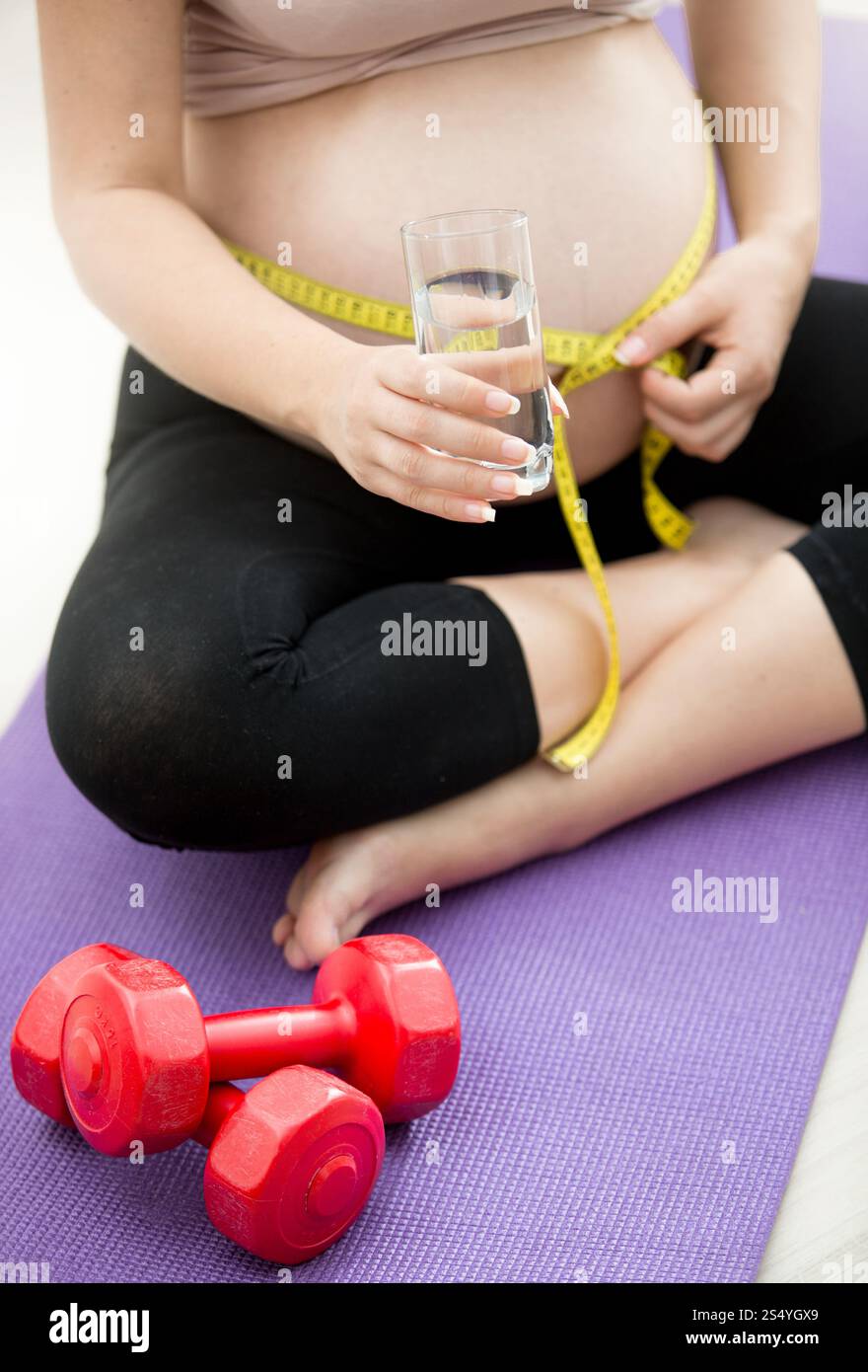 Gros plan d'une femme enceinte mesurant le ventre avec un ruban à mesurer sur un tapis de fitness et tenant un verre d'eau Banque D'Images
