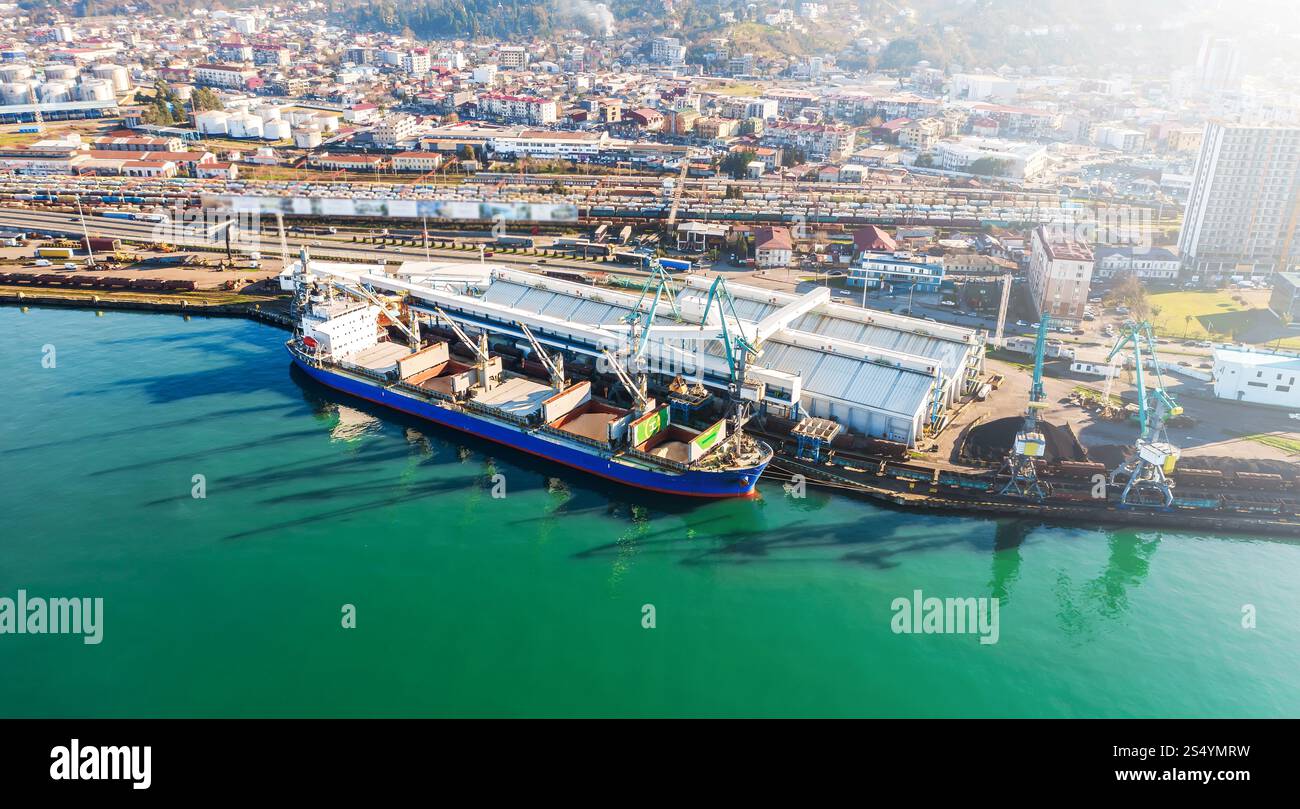Navire cargo amarré dans un port de ville animé avec des cales chargées et des ponts roulants, reflétant les routes commerciales de la chaîne d'approvisionnement mondiale et le commerce maritime moderne. Banque D'Images