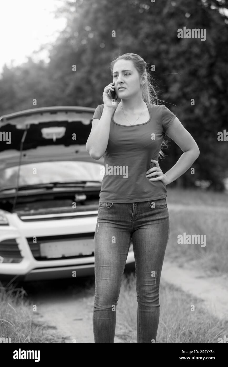 Portrait noir et blanc d'une jeune femme en colère debout à la voiture cassée et parlant par téléphone portable Banque D'Images