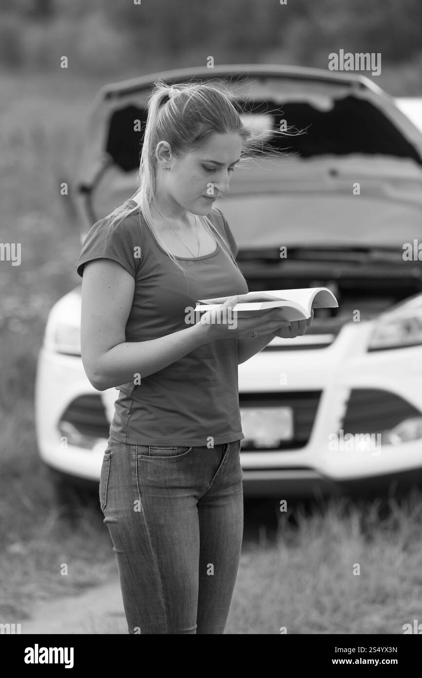 Image en noir et blanc d'une femme déprimée regardant sa voiture cassée et lisant le manuel du propriétaire Banque D'Images