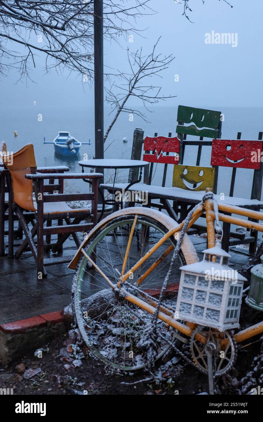 Un vélo jaune vif couvert de neige fraîche repose près d'un café au bord du lac par un matin d'hiver tranquille. Chaises colorées au visage souriant Banque D'Images