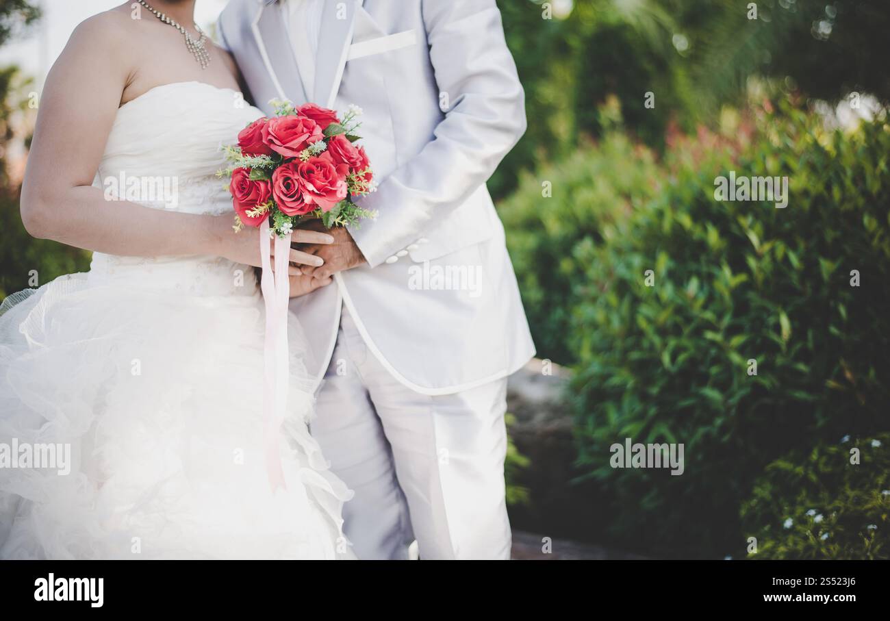 Mariée tient un bouquet de rose rouge de mariage dans les mains, les câlins du marié. Mariée tient un bouquet de rose rouge de mariage dans les mains, le marié embrasse sa mariée ensemble. Banque D'Images