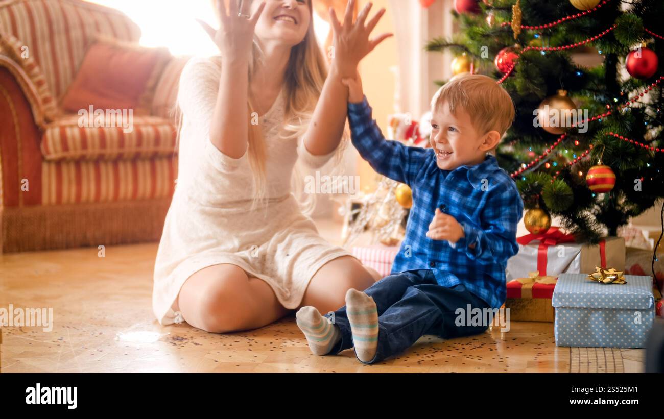 Joyeux garçon en bas âge riant jetant des confettis colorés avec la mère le matin de Noël. Portrait de joyeux garçon en bas âge riant jetant coloré Banque D'Images