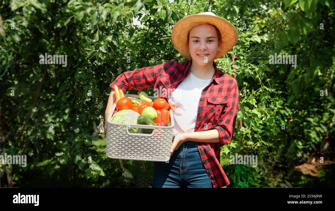 Fille souriante dans le chapeau marchant à arden avec la récolte dans une grande boîte. Légumes biologiques frais et mûrs en boîte. Concept de petite entreprise et de croissance organique Banque D'Images