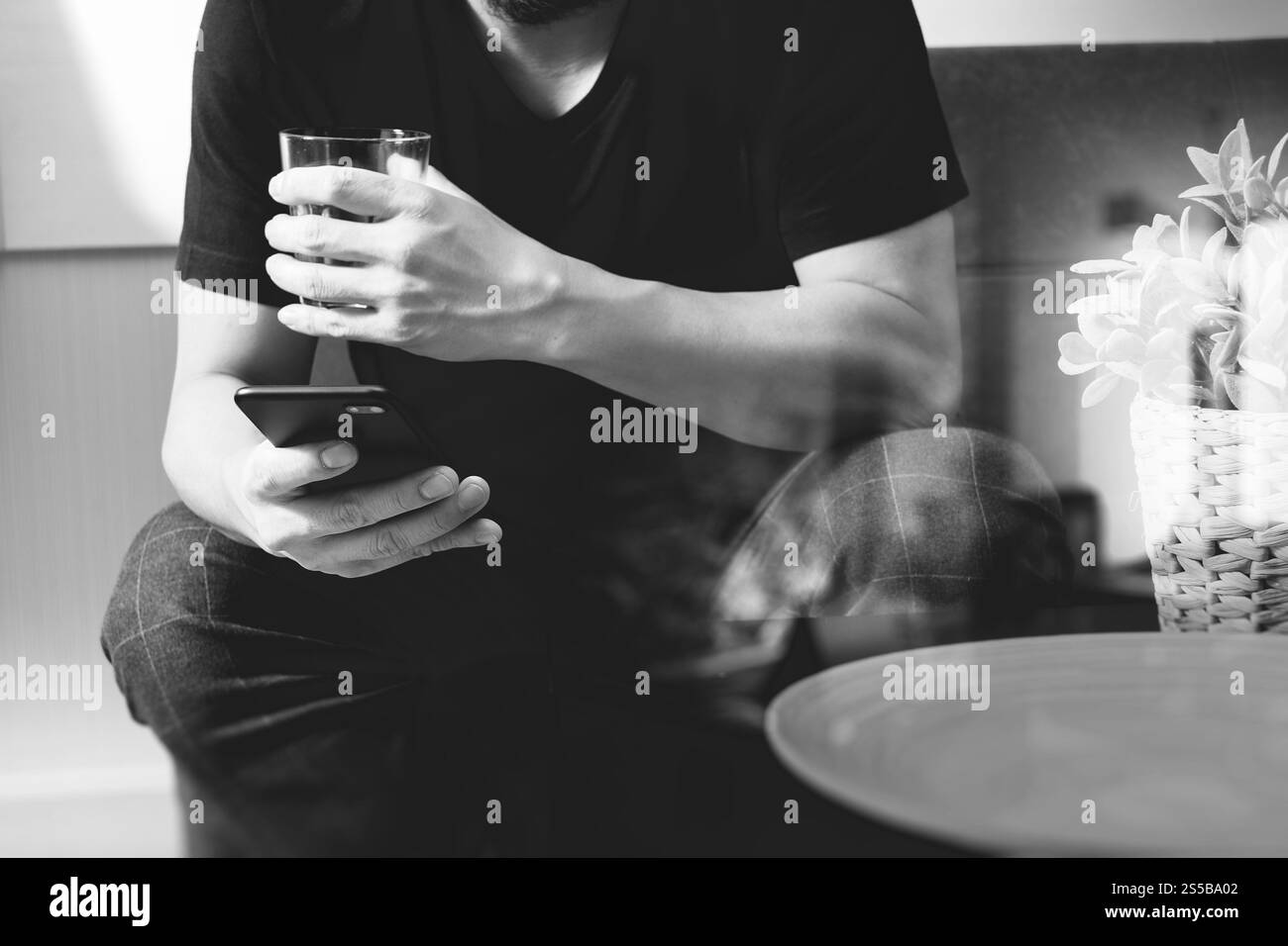 main d'homme designer avec verre d'eau en utilisant le phon intelligent pour les paiements mobiles shopping en ligne, canal omni, assis sur le canapé dans le salon, vase en rotin Banque D'Images