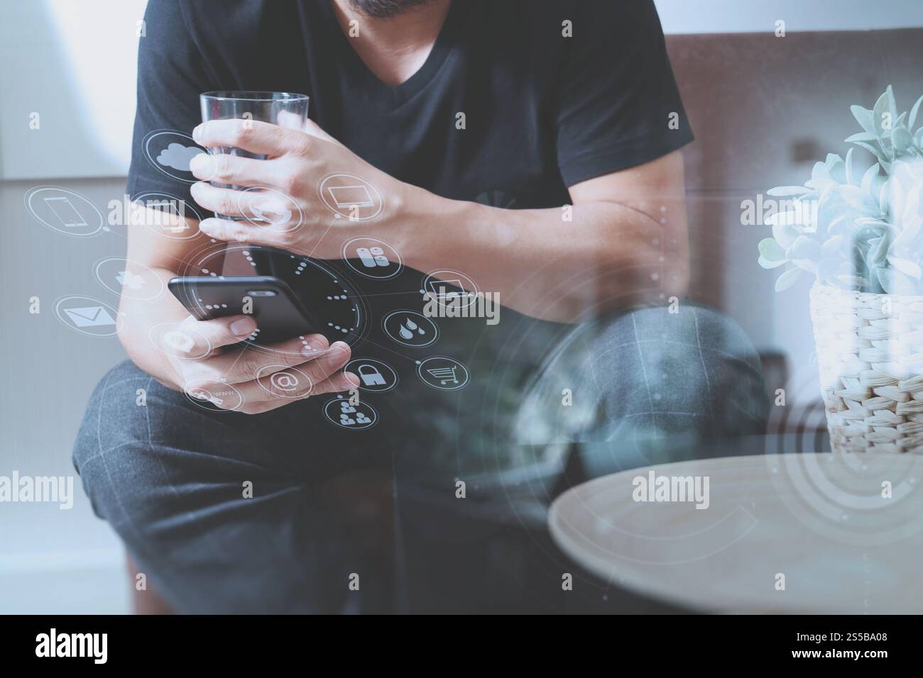 main d'homme designer avec verre d'eau en utilisant le phon intelligent pour les paiements mobiles shopping en ligne, canal omni, assis sur le canapé dans le salon, vase en rotin Banque D'Images