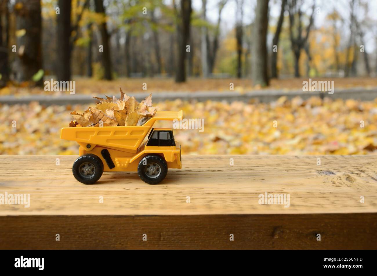 Un petit camion jaune jouet est chargé de feuilles jaunes tombées. La voiture se tient sur une surface en bois sur un fond d'un parc d'automne flou. Banque D'Images