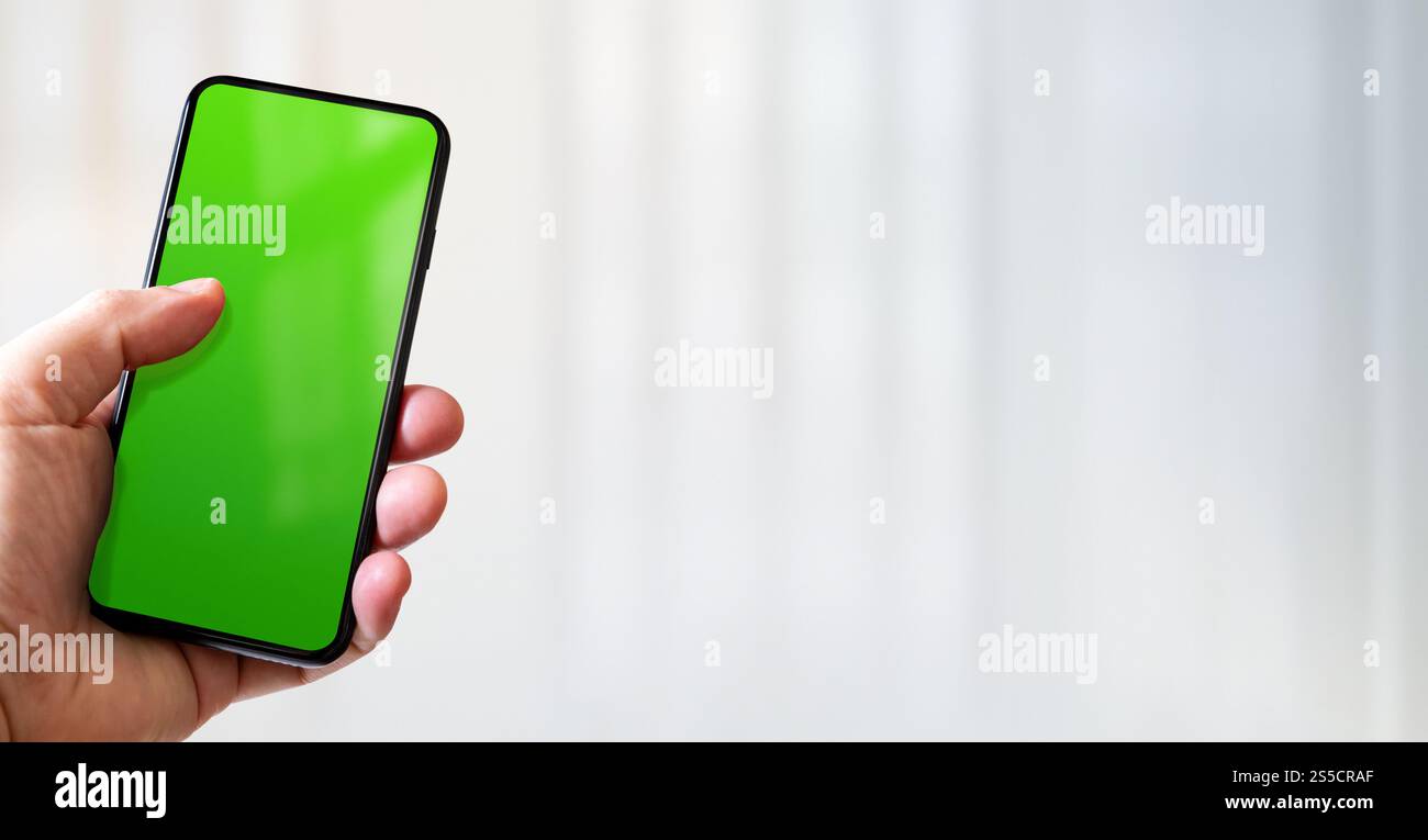 Main tenant un smartphone avec écran vert vierge. Fond blanc de bureau. Bannière horizontale. Main tenant un smartphone avec écran vert vierge. Banque D'Images