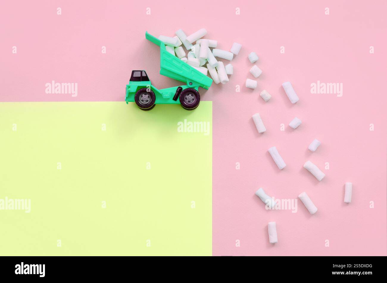 Green Little Toy dump truck jette des morceaux de guimauve de son dos soulevé sur un fond jaune pastel et corail. Vue de dessus minimale à plat. Vidage Banque D'Images