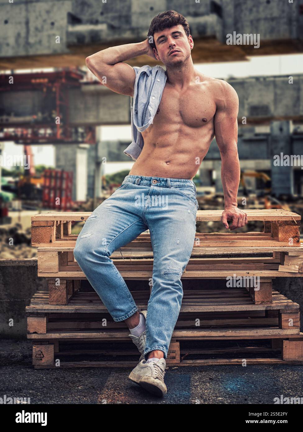 Un modèle masculin musclé se tient décontracté sur des palettes en bois sur un chantier de construction, mettant en valeur son physique tonique tout en portant un Jean bleu clair et des baskets. Le cadre est animé par l'activité. Banque D'Images
