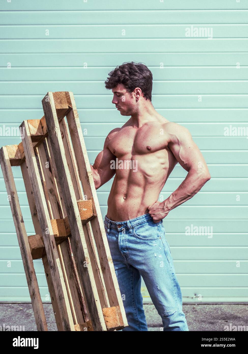 Un jeune homme musclé se tient dehors, examinant une palette en bois avec une main sur sa hanche. Il porte un t-shirt simple et un Jean, mettant en valeur un physique ajusté sur un fond clair. Banque D'Images