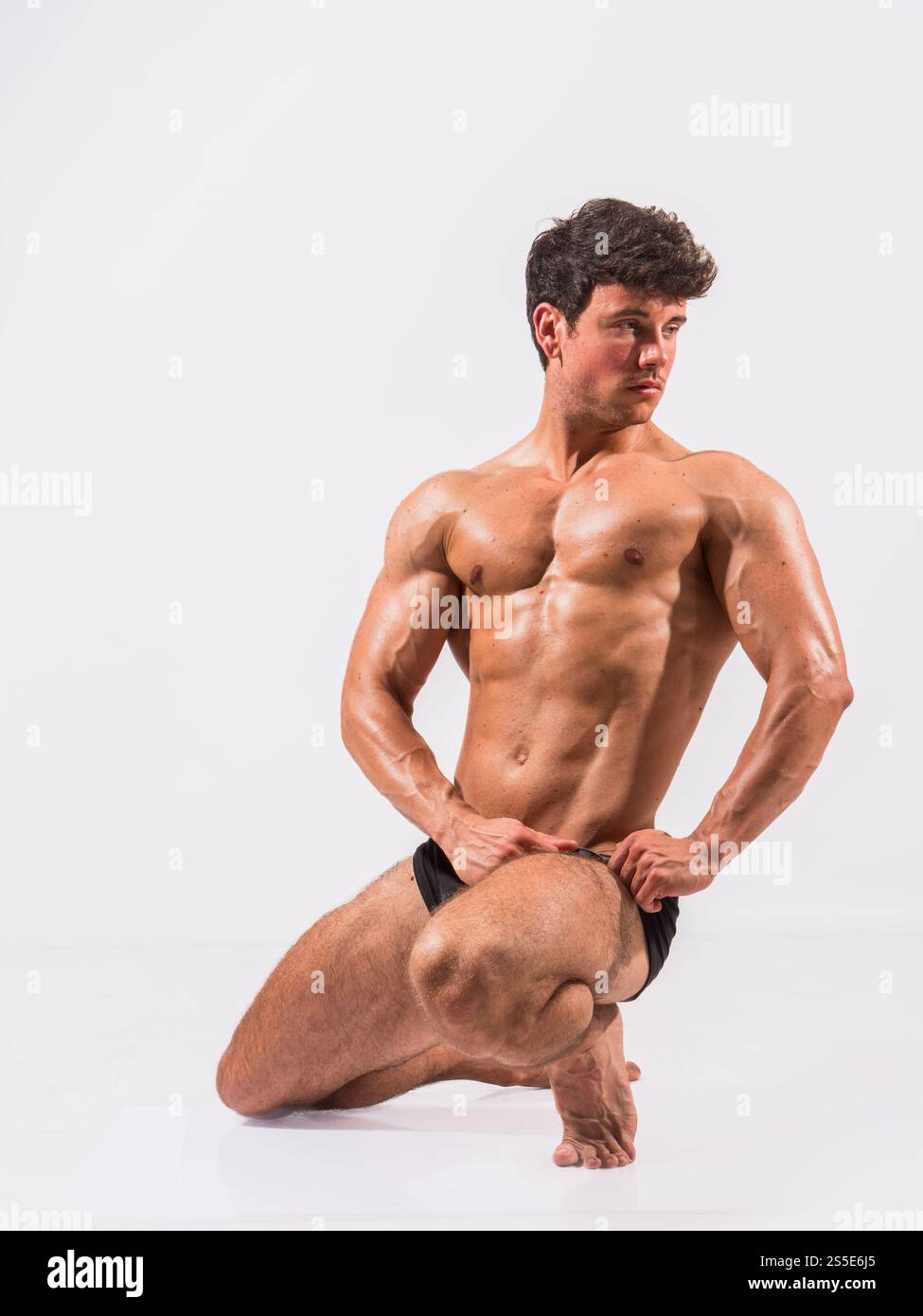 Un modèle de fitness masculin s'agenouille dans une pose forte, soulignant son physique musculaire. Le modèle affiche confiance et force sur un fond Uni, mettant l'accent sur l'art de sculpter le corps. Banque D'Images