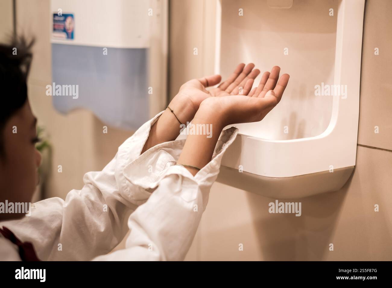 Garçon asiatique en chemise à manches longues séchant ses mains en utilisant le sèche-mains dans les toilettes publiques Banque D'Images