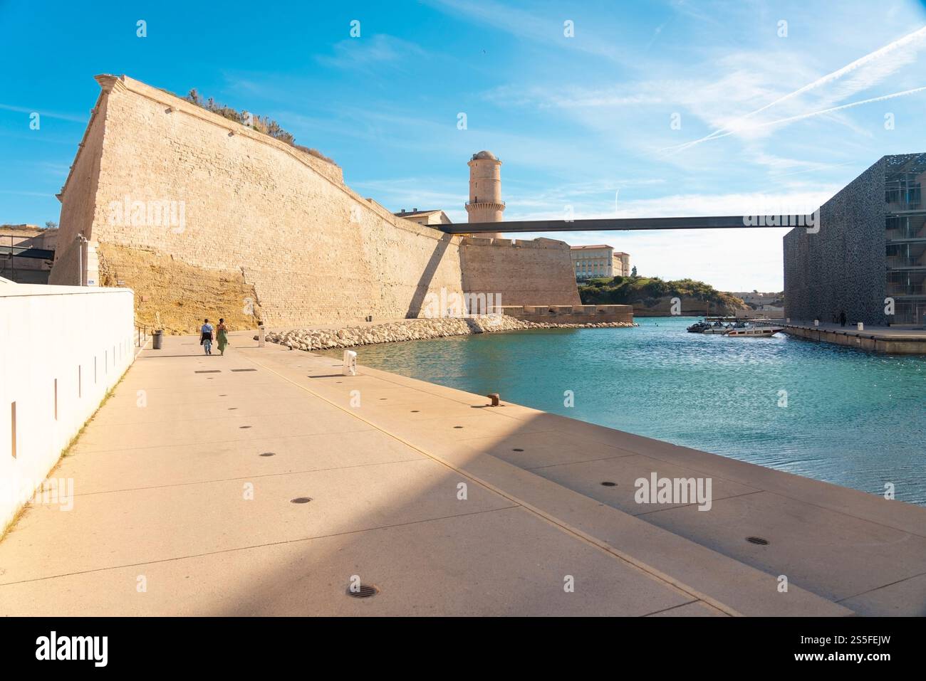 Une vue panoramique d'un fort historique du mur de Saint Jean à côté d'un plan d'eau serein méditerannean avec des gens se promenant le long de la passerelle adjacente sous un ciel bleu clair, Marseille, France Banque D'Images