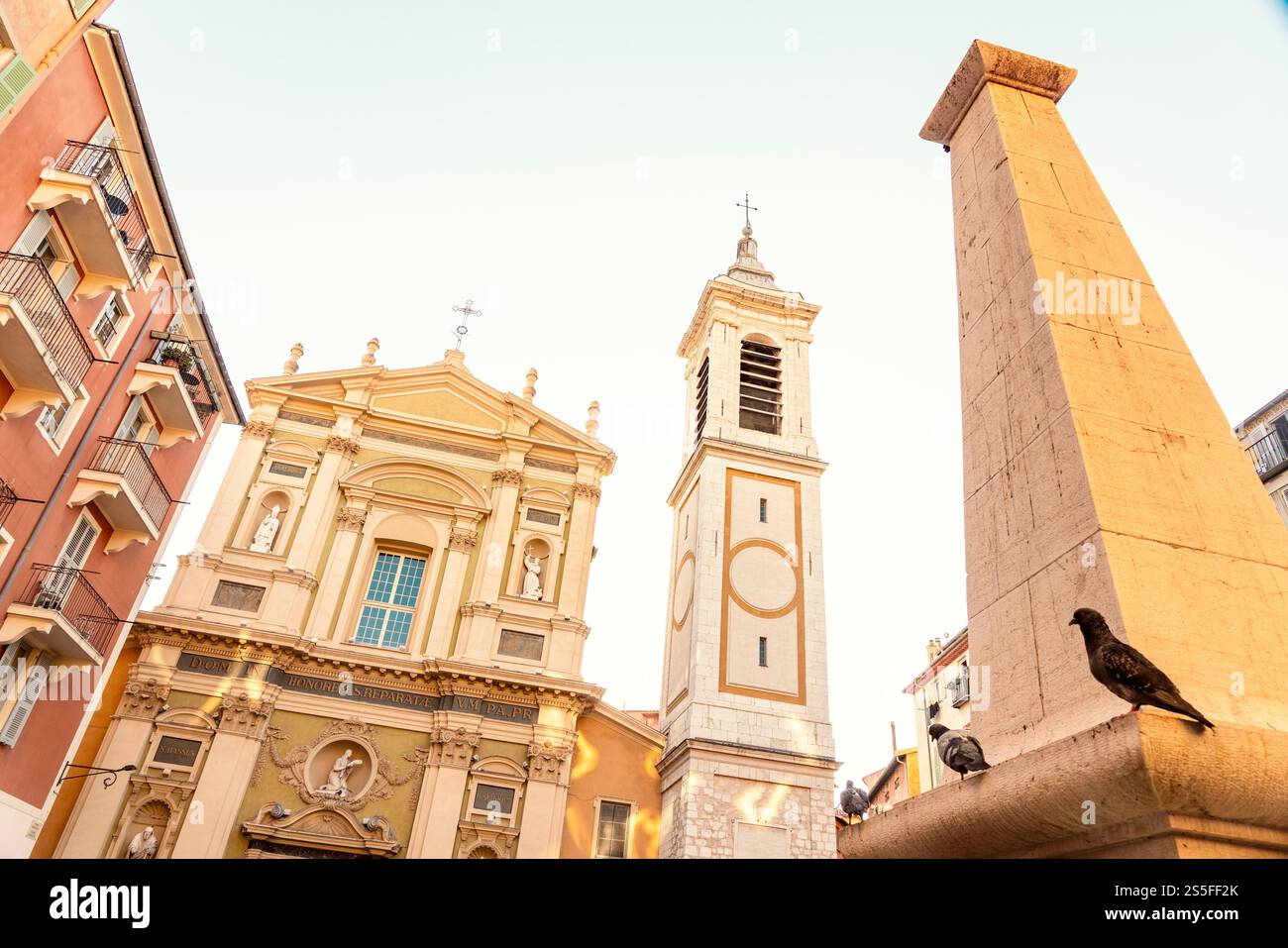 Un pigeon perche sur un rebord ensoleillé avec une église baroque et un obélisque en arrière-plan contre un ciel bleu clair, Nice, France Banque D'Images Un pigeon perche sur un rebord ensoleillé avec une église baroque et un obélisque en arrière-plan contre un ciel bleu clair, Nice, France Banque D'Images
