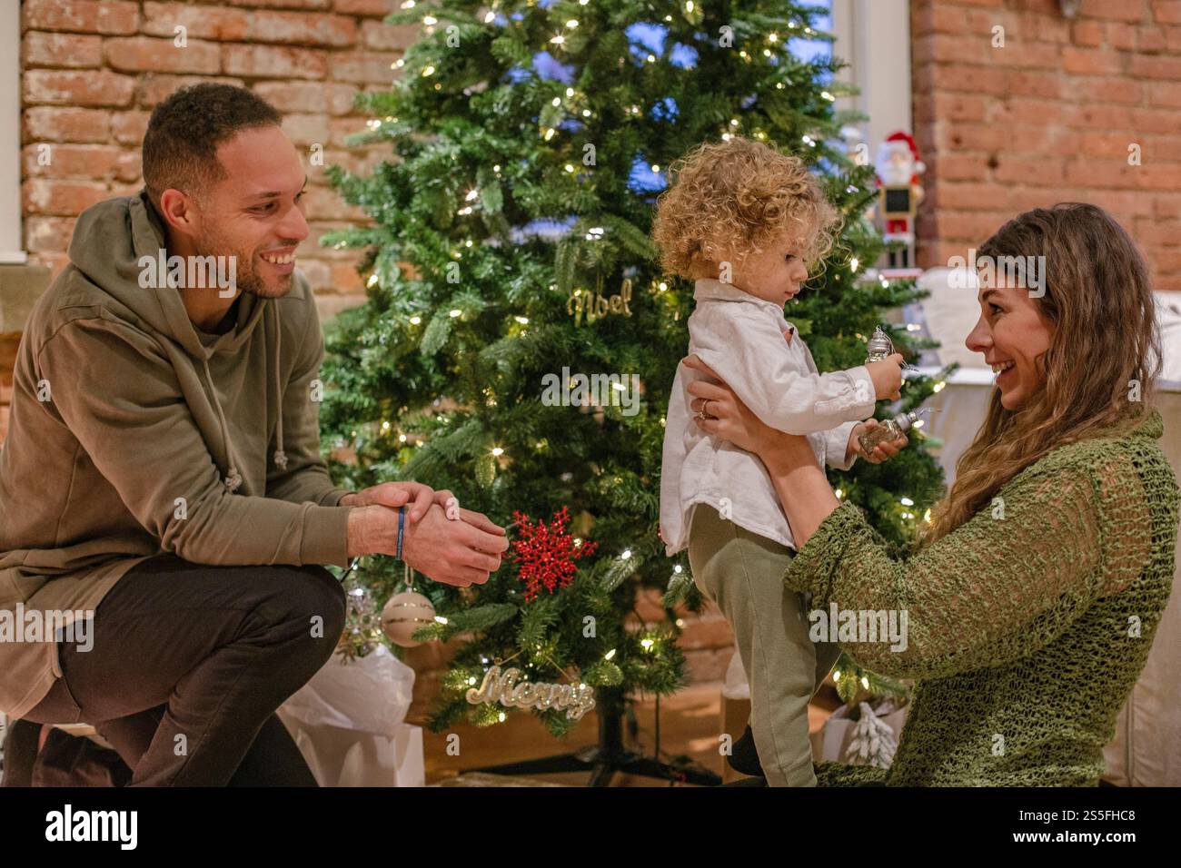 Une famille heureuse avec un enfant en bas âge décorant un arbre de Noël dans un cadre de maison confortable, Ohio, États-Unis Banque D'Images