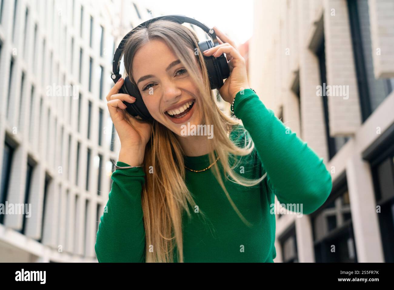 Femme souriante dans un top vert aime la musique sur ses écouteurs dans un cadre urbain, Berlin, Allemagne Banque D'Images