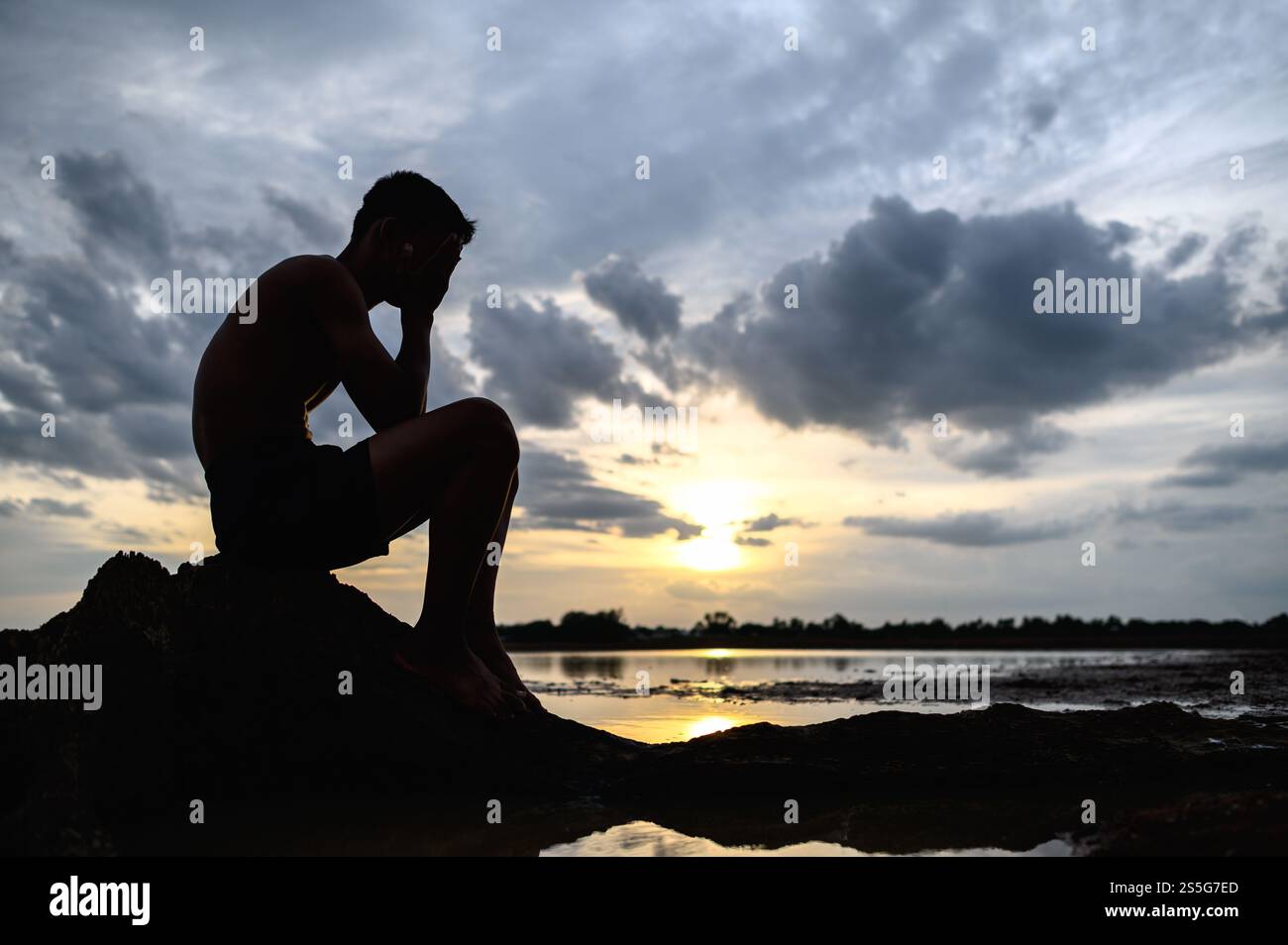 Un homme s'assit les genoux, en tenant ses mains sur le visage à la base de l'arbre et il y a de l'eau autour. Banque D'Images