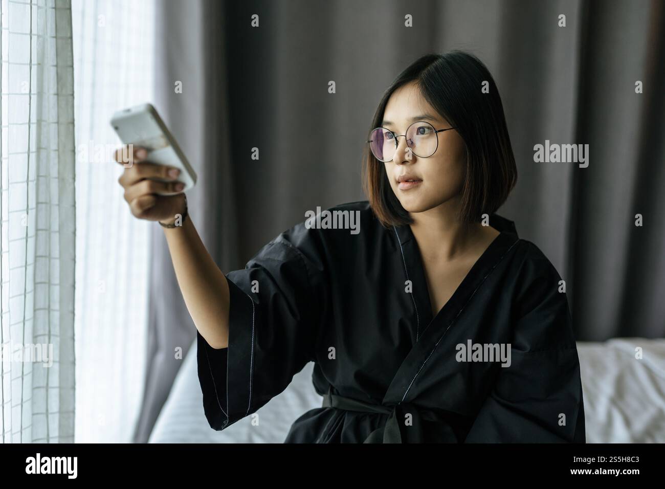Femme portant un manteau noir, portant des lunettes, assise, appuyant sur la télécommande pour allumer l'air. Banque D'Images
