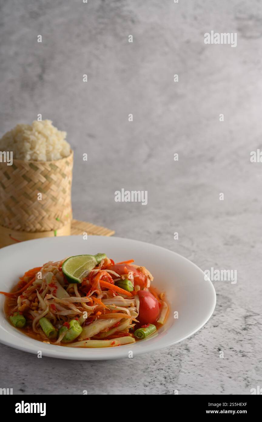 Salade de papaye thaïlandaise dans une assiette blanche avec riz collant dans un panier en osier en bambou. Banque D'Images