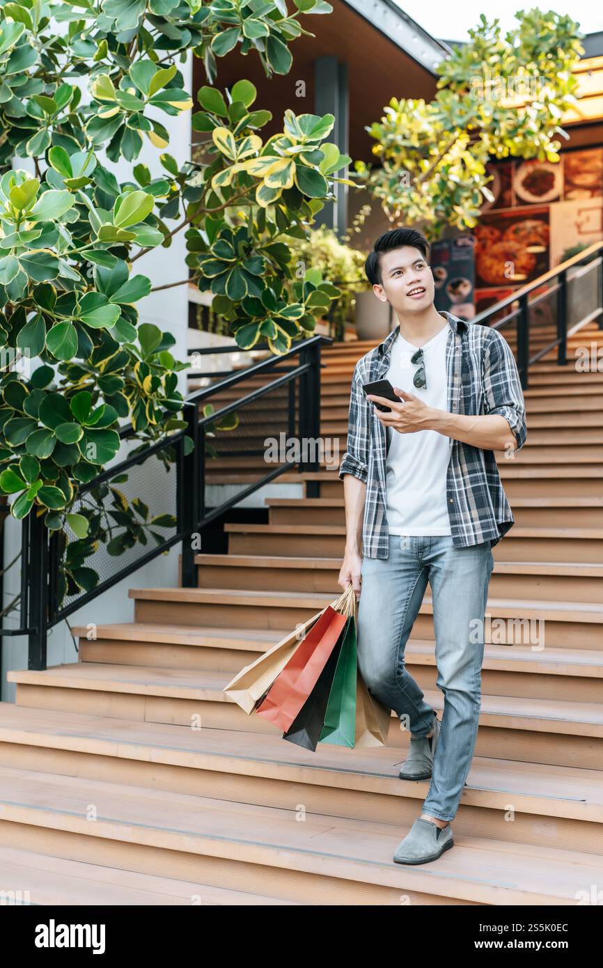 Portrait jeune homme beau tenant un sac en papier et utiliser un smartphone sur l'escalier du centre commercial, style de vie et nouveau concept normal, espace de copie Banque D'Images