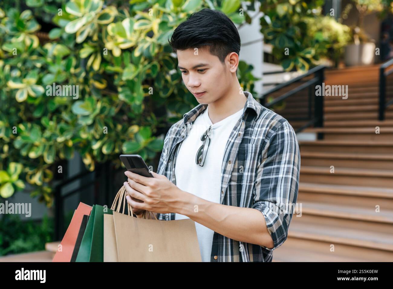 Portrait jeune homme beau tenant un sac en papier et utiliser un smartphone sur l'escalier du centre commercial, style de vie et nouveau concept normal, espace de copie Banque D'Images